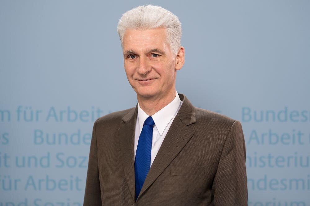 Porträtfoto von Dr. Rolf Schmachtenberg, Beamteter Staatssekretär im Bundesministerium für Arbeit und Soziales. Herr Dr. Schmachtenberg trägt einem braunen Anzug mit blauer Krawatte und steht vor einem hellblauen Hintergrund, auf dem Teile des der Worte „Bundesministerium für Arbeit und Soziales“ zu sehen sind. – 