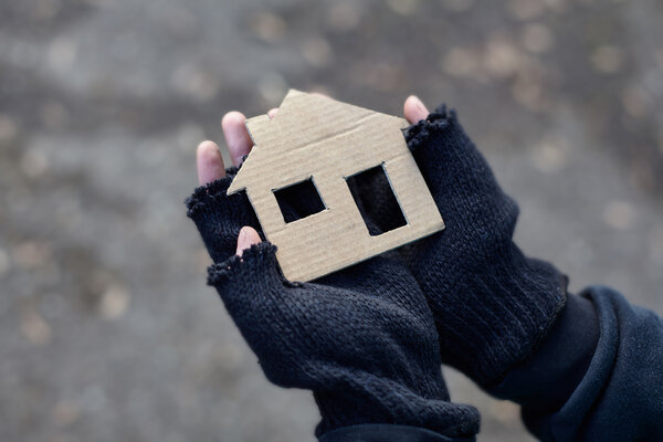 Zwei Hände in schwarzen Handschuhen, die behutsam ein Haus aus Pappe halten. – 