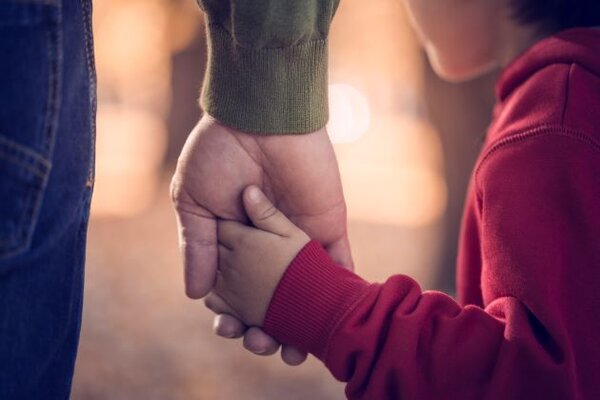 Das Foto ist ein Ausschnitt von einer Hand einer erwachsenen Person, die die Hand eines Kindes hält. Das Kint trägt eine rote Jacke. Die erwachsende Person eine dunkelblaue Hose und ein grünes Hemd. – 